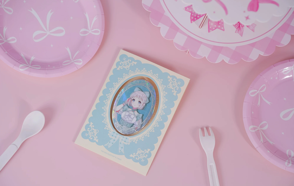 The Lilja Katsuragi badge on its backing card, photographed on a pink table with paper plate and cutlery props